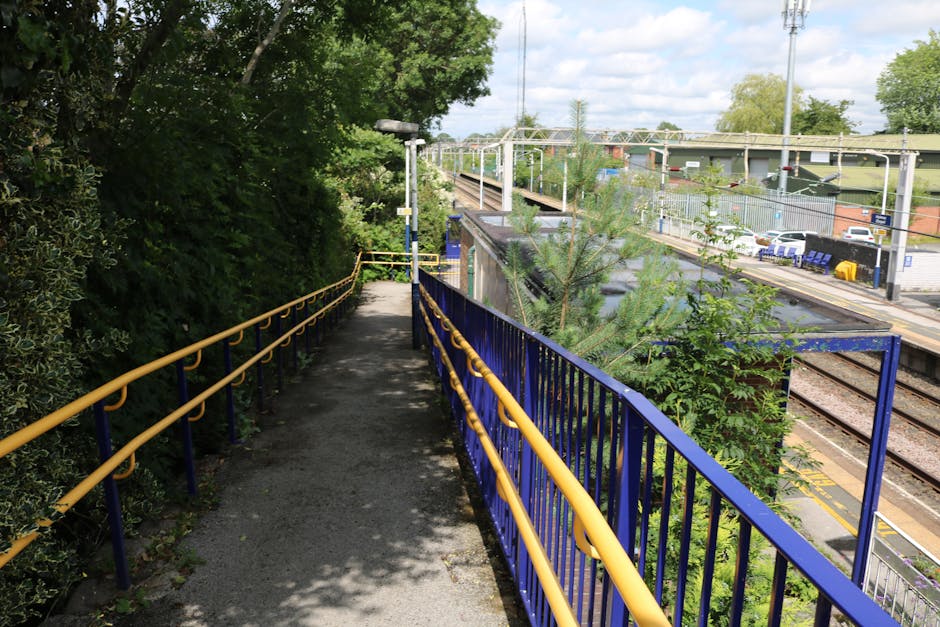 A narrow outdoor footpath with a black asphalt surface, bordered by yellow handrails on the left and blue railings on the right, runs alongside a railway track at a train station. The path is shaded by green trees and bushes on the left side, with sunlight filtering through the foliage. In the distance, there is a small evergreen tree growing close to the railings. Beyond the railings, the railway platform features a row of parked cars, a shelter, and train tracks that extend into the horizon. Overhead electrical lines and metal supports are visible above the platform, with a partly cloudy sky overhead. This environment appears to be part of a home relocation process, with the surrounding area suitable for furniture transport and the loading process towards the station managed by Man with Van Noak Hill, supporting efficient removals and moving logistics.