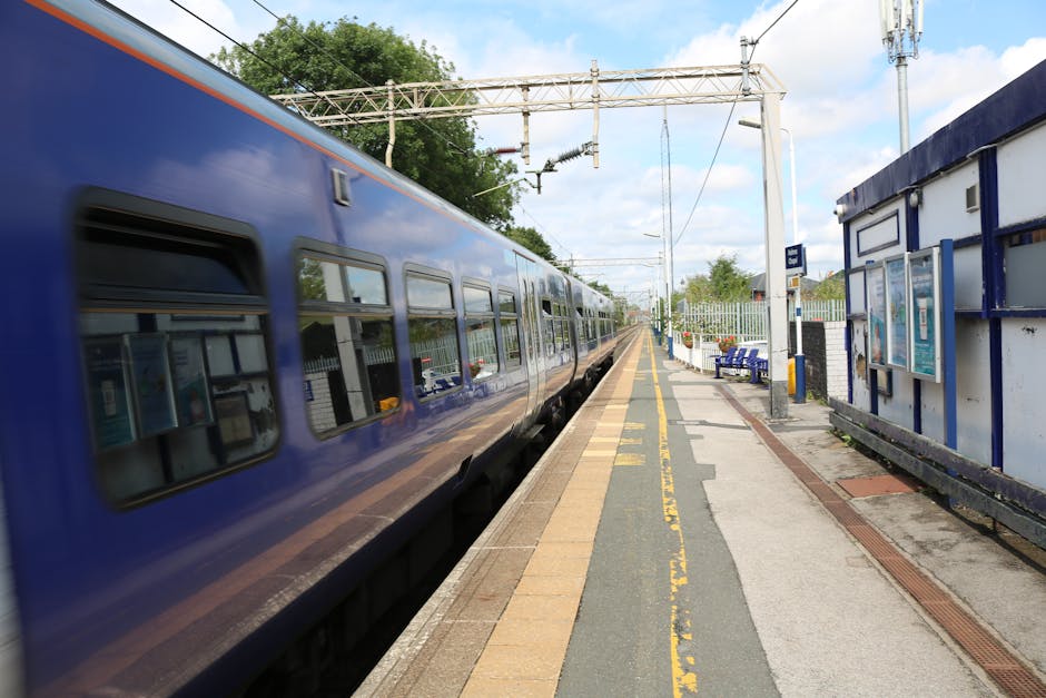 A blue passenger train is stationary at a platform, with closed windows reflecting the outdoor environment. The platform is paved with a mix of concrete and brick, featuring yellow safety lines along the edge. To the right, there is a small shelter with advertising posters and a blue painted wall. Overhead, electrical wires and a metal gantry support the railway's electrification system. Several blue benches are positioned on the platform, some with personal belongings, indicating preparations for a home relocation or furniture transport process. Part of a manual trolley is visible on the back of the platform, suggesting the loading of packing boxes or furniture. The scene is outdoors, with a partly cloudy sky and green trees in the background, capturing a moment in the logistics of house removals or moving services conducted by Man with Van Noak Hill.