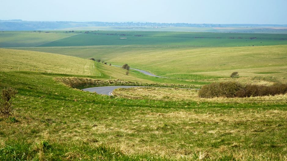 A winding country road with a smooth asphalt surface featuring white dashed navigation lines, curving gently through rolling green hills under an overcast sky. The roadside verges are lined with grass