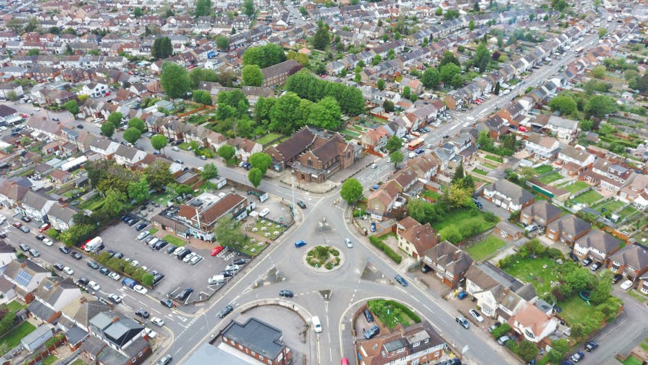 An aerial view of a residential neighbourhood in Noak Hill showing a central roundabout with multiple roads branching outward. Surrounding the roundabout are rows of terraced and semi-detached houses with brick facades and tiled roofs, interspersed with green trees, gardens, and small front yards. Near the roads, there are parked cars along the curb and within designated parking areas. The scene includes a mix of blue, white, and grey vehicles, with some properties having visible fences and patios. Man with Van Noak Hill is involved in a home relocation process, likely utilizing these streets for furniture transport and loading belongings onto a van parked nearby, which can be inferred from the context of quick, same-day removals services. The area appears well-lit, with natural daylight illuminating the scene, capturing the organized logistics of residential moving or packing and moving activities.