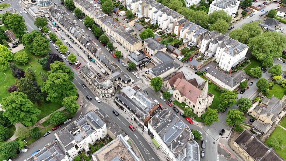 An aerial view of a residential street in Noak Hill showing a variety of houses, including terraced, semi-detached, and detached properties, with rooftops made of slate and tile. The street is lined with numerous parked cars along both sides and is surrounded by abundant green trees and small parks, providing a tranquil neighbourhood environment. In the foreground, a church with a pointed steeple and light-colored stone walls is visible, situated among other buildings. The scene includes a section of pavement and a narrow alleyway adjacent to the houses. The image captures the typical urban landscape where home relocation and furniture transport activities by Man with Van Noak Hill could take place, especially around the central area near the church and residential frontages, with pathways and open spaces suitable for loading and unloading during removals.