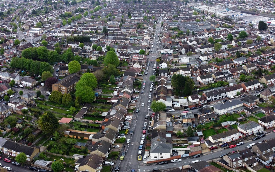 An aerial view of a residential neighbourhood in Noak Hill showing a central roundabout with multiple roads branching outward. Surrounding the roundabout are rows of terraced and semi-detached houses with brick facades and tiled roofs, interspersed with green trees, gardens, and small front yards. Near the roads, there are parked cars along the curb and within designated parking areas. The scene includes a mix of blue, white, and grey vehicles, with some properties having visible fences and patios. Man with Van Noak Hill is involved in a home relocation process, likely utilizing these streets for furniture transport and loading belongings onto a van parked nearby, which can be inferred from the context of quick, same-day removals services. The area appears well-lit, with natural daylight illuminating the scene, capturing the organized logistics of residential moving or packing and moving activities.
