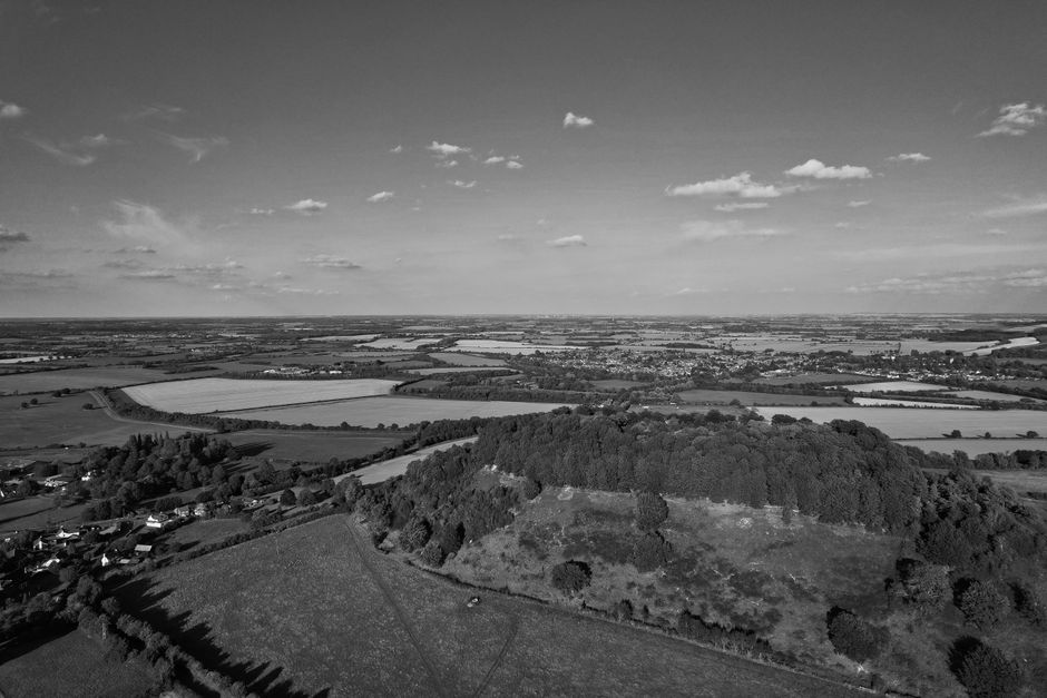 A black-and-white aerial photograph depicting a rural landscape with expansive fields, a small cluster of houses in the lower left corner, and a large wooded hill in the foreground. The open fields are separated by hedgerows and narrow roads, extending towards the horizon where the sky, dotted with scattered clouds, meets distant farmland. The scene captures a serene countryside environment, with no visible furniture, packaging, or moving equipment. This image could relate to house removals or home relocation services by [COMPANY_NAME], illustrating the rural setting where logistics may involve planning for transportation routes through countryside areas, such as during a furniture transport or packing and moving process.