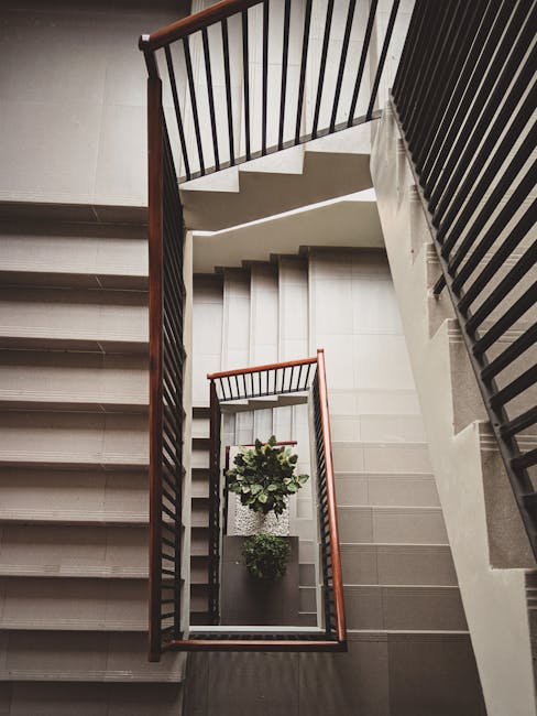 A top-down view of a multi-level staircase in an interior building, showing a central rectangular landing with tiled flooring. The staircase has wooden handrails with vertical black metal balustrades, and white steps leading both upwards and downwards. Two small potted plants are positioned on the landing near the wall, and the surrounding walls are painted in a light neutral colour. The area is well-lit, highlighting the clean, orderly appearance of the staircase, which is part of a residential property. This image reflects the type of location where a household move or relocation by Man with Van Noak Hill could involve furniture transport and packing services through stairway-only access, aligning with house removals and moving logistics within such flats in Noak Hill.