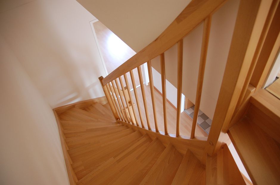 A top-down view of a multi-level staircase in an interior building, showing a central rectangular landing with tiled flooring. The staircase has wooden handrails with vertical black metal balustrades, and white steps leading both upwards and downwards. Two small potted plants are positioned on the landing near the wall, and the surrounding walls are painted in a light neutral colour. The area is well-lit, highlighting the clean, orderly appearance of the staircase, which is part of a residential property. This image reflects the type of location where a household move or relocation by Man with Van Noak Hill could involve furniture transport and packing services through stairway-only access, aligning with house removals and moving logistics within such flats in Noak Hill.